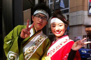 黄緑色の衣装を着た男性と、赤色の衣装を着た女性が並び笑顔でピースサインをしている写真