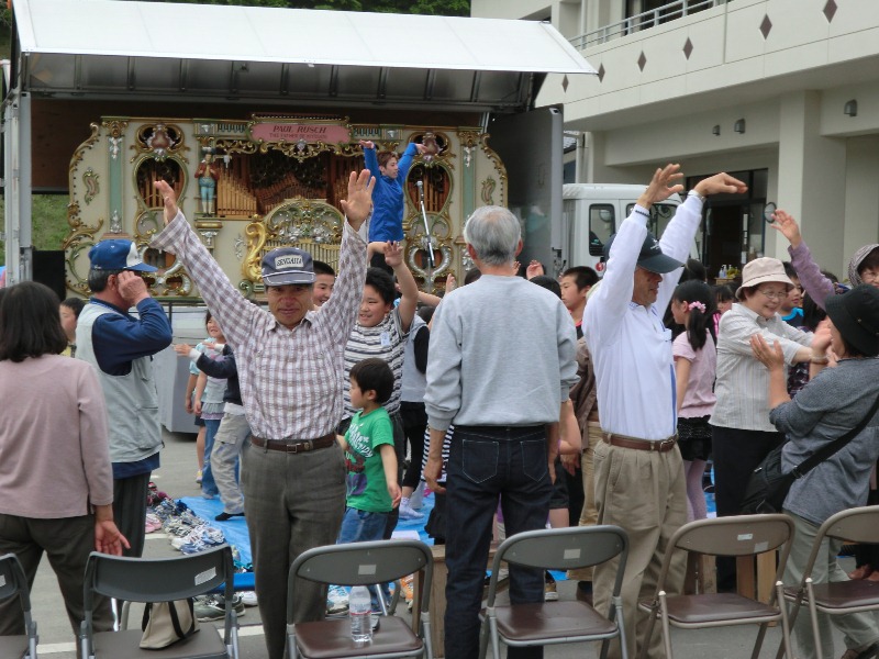 前方に世界最大のオルゴールが置かれた会場で、大人も子どもも両手を上に挙げてバレエの動きをまねて体を動かし、楽しそうに踊っている様子を写したバレエ体験の写真