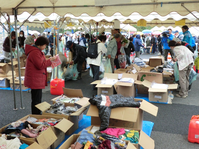 テントの下に多数の段ボール箱が並べられ、その中に衣類などの品物が入っていて、大勢の人々が品物を見たり手に取ったりしている様子を写した写真
