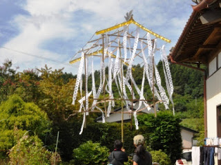 竹の骨組みに白い紙の飾りが吊るされた伝統的な装飾が庭先に設置され、男性たちがそれを眺めている写真