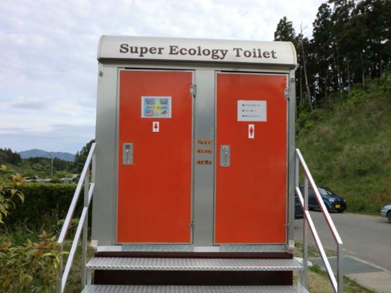 階段を上って入る構造の「Super Ecology Toilet」と書かれたシルバーの箱型で、正面にオレンジ色の扉が2つ並ぶバイオトイレを写した写真