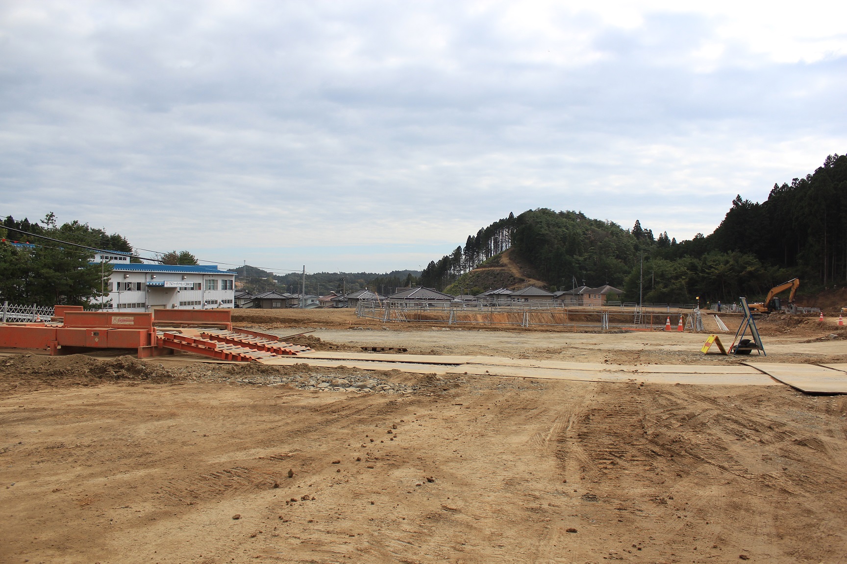 曇り空のもと、茶色い土が広がる造成地では、手前にオレンジ色のタイヤ洗浄設備が設置され、奥には住宅と木々に囲まれた小高い山が見え、重機による整地作業が続けられている写真