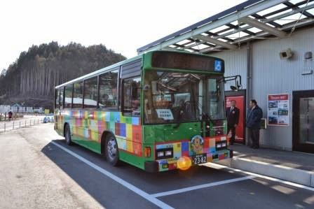 デザインされたカラフルな四角形の模様がラッピングされた、緑色のバスが停留所に停車している様子を写した写真