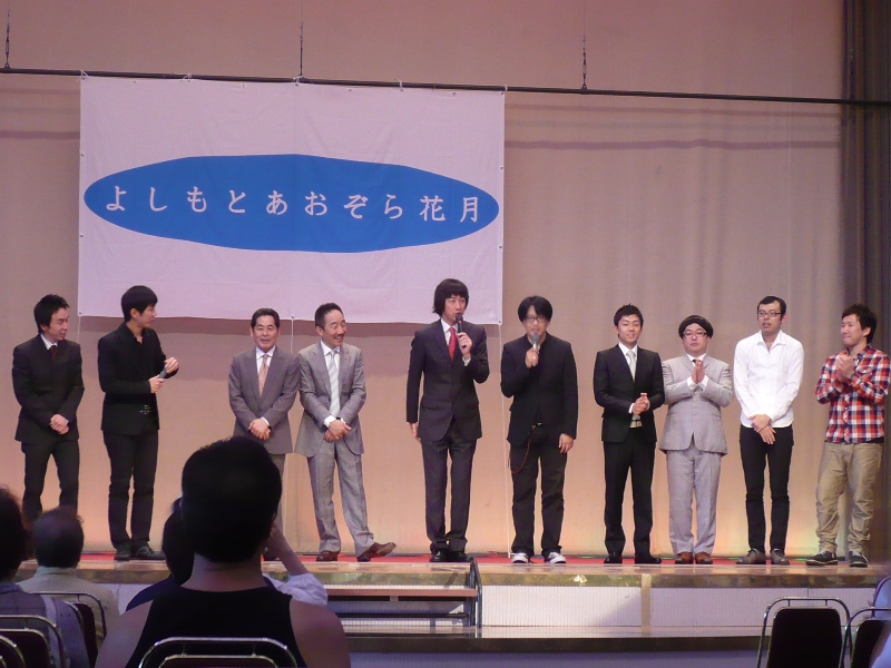 「よしもとあおぞら花月」と書かれた大きな横断幕の前で、吉本の芸人の方々が舞台に立ち、中央に立つ2人がマイクを持って話をしている様子を写した写真