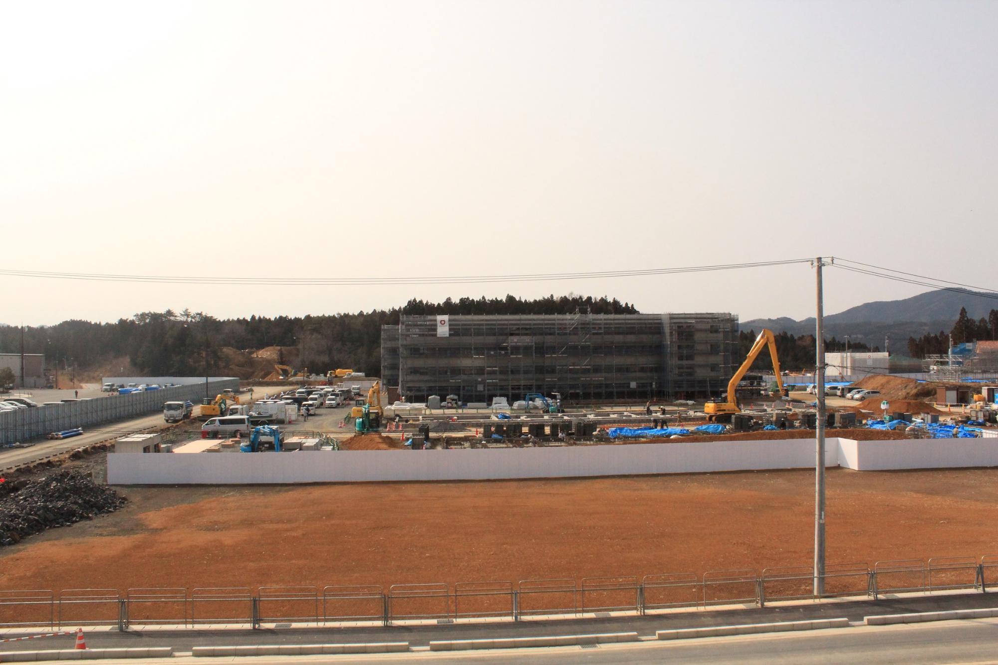 重機や作業車が行き交う広大な建設現場で、足場に覆われた建物が中央にそびえ、周囲には資材や車両が整然と並び、前景には赤茶けた整地済みの土地が広がっている写真