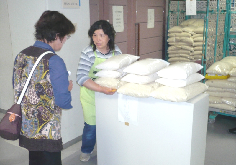 複数の米袋が積まれたカウンター越しに、黄緑色のエプロンを着た女性が訪れた女性とやり取りしており、背景にも棚や台車に積まれた米袋が見える、自立支援米提供の様子の写真