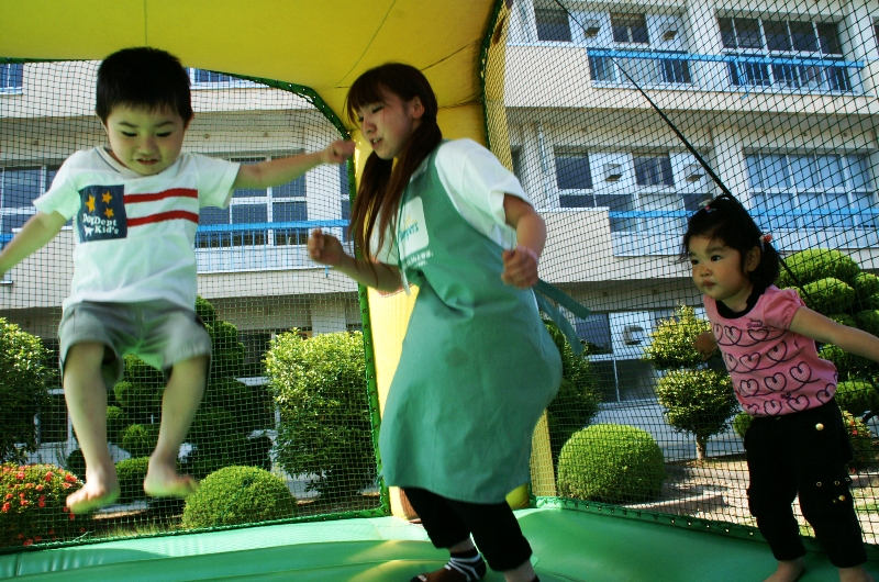 屋外に設置された黄色い屋根のエアートランポリンの中で、エプロン姿のお姉さんと幼い男の子と女の子が飛び跳ねて遊んでいる様子を写した写真