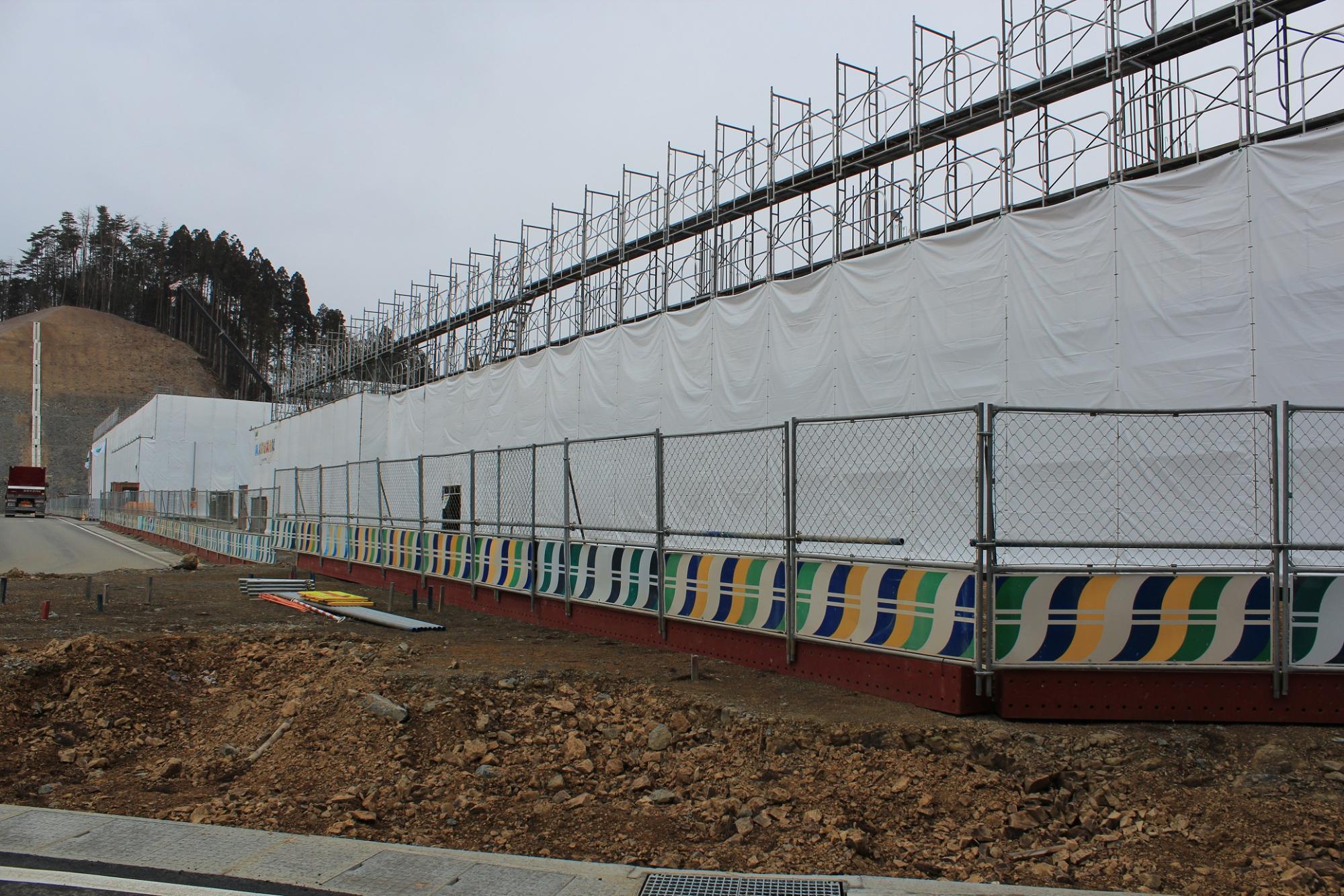 白いシートで覆われた高い足場が長く連なり、その手前にはカラフルな模様の仮囲いが設置され、足元には掘り起こされた土や岩、散らばった工具類が見える山あいの建設現場の様子を捉えた写真