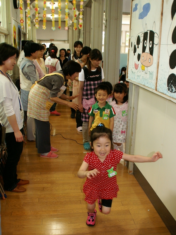 エプロン姿の保育士や関係者が見守る中、保育室から笑顔で走りだしてくる女の子と、順番に廊下へ出てくる笑顔の子どもたちの様子を写した写真