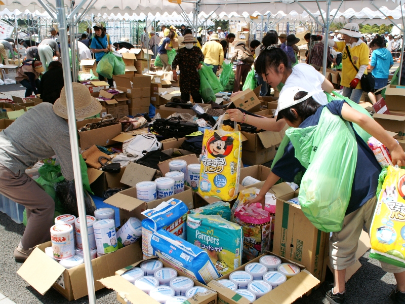 テントの下に段ボール箱がたくさん並び、ミルク缶や紙おむつ、日用品などの品物を多くの人が選びながら見ているフリーマーケットの様子を写した写真