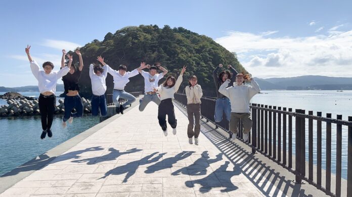海に囲まれた堤防の上で、9人の若者が快晴の青空の下、島を背にして元気よくジャンプしており、それぞれが両手を上げたり笑顔を見せたりしており、影が足元にくっきり映っている活気ある一瞬を捉えた写真