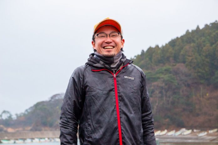 木々と岸辺に船や建物が遠目に見える水辺に立ち、オレンジ色のキャップをかぶり、黒いジャケットを着て笑顔の後藤伸弥さんの写真