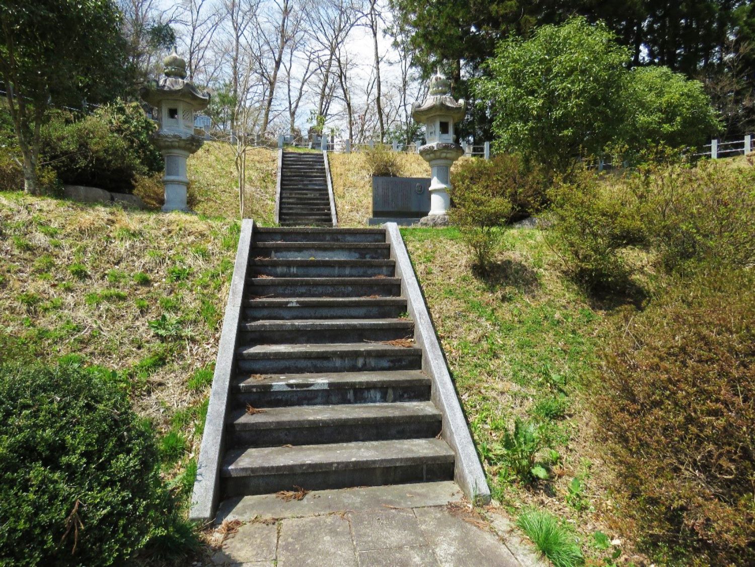 周囲に木々や低木が茂る神聖な雰囲気の丘に石階段がまっすぐ伸びており、階段の上には左右対称に石灯籠が立ち、右側の石灯籠の奥には記念碑が設置されている写真