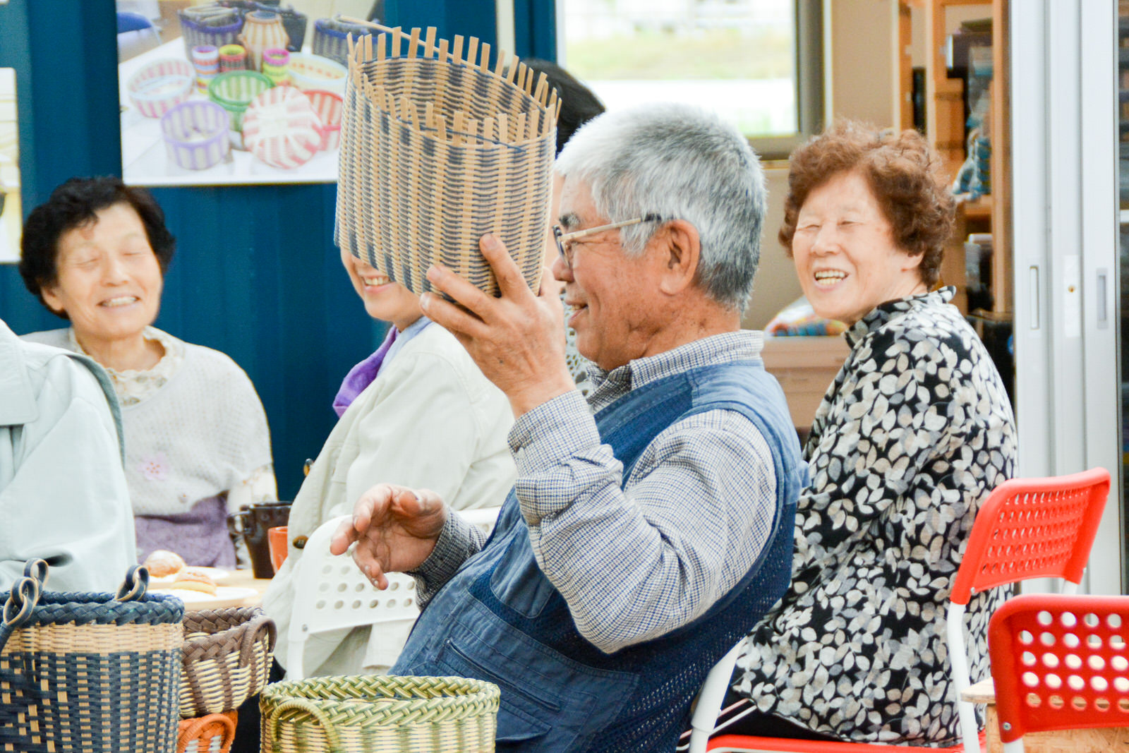 年配の男性が編みかごを手に取りながら笑い、周囲の高齢者も笑顔で楽しそうにしている写真