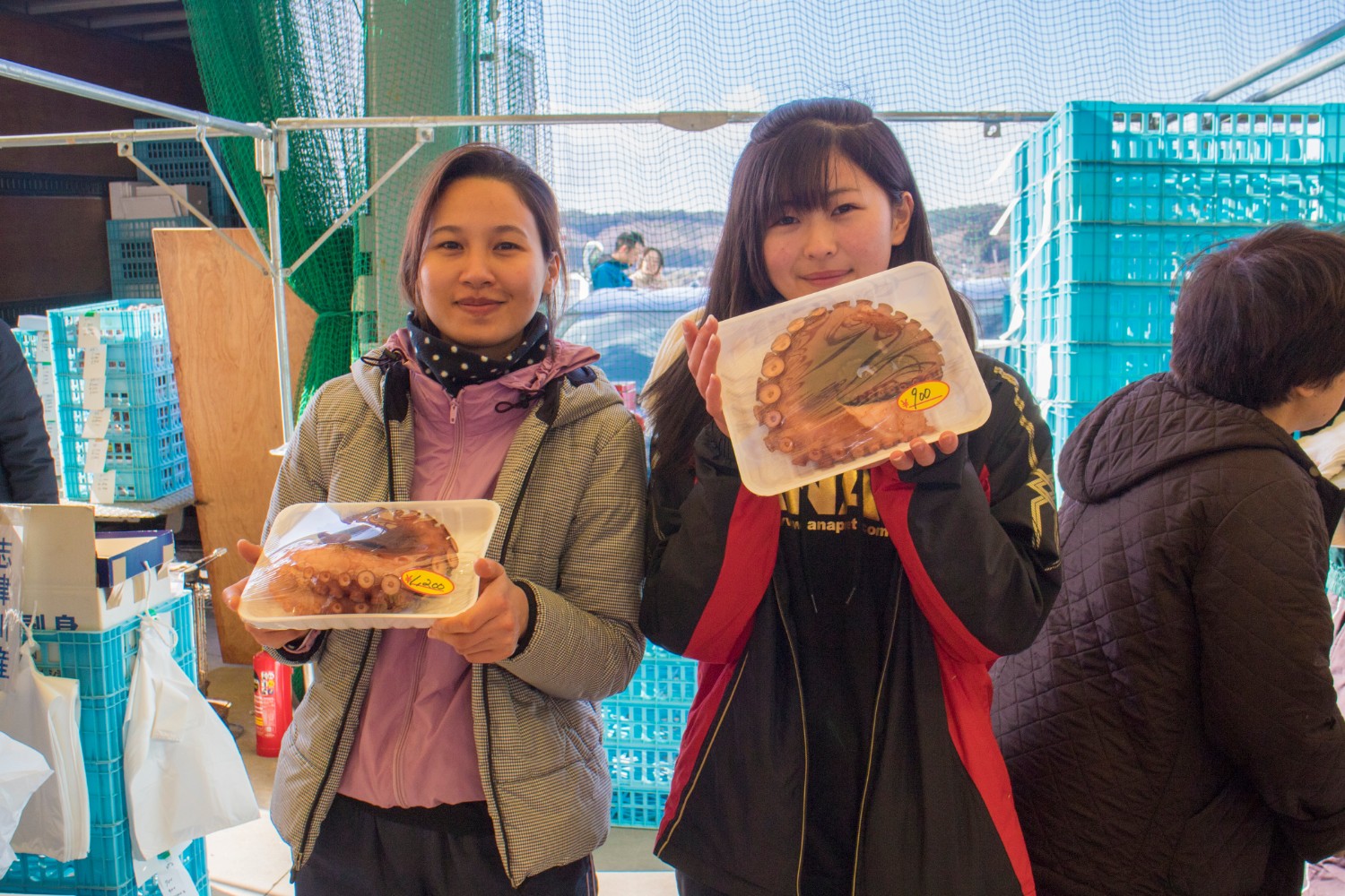 屋外の市場の一角で、若い女性二人がそれぞれ透明フィルムで包まれた大きなゆでだこのパックを両手で持ち笑顔で写っている写真