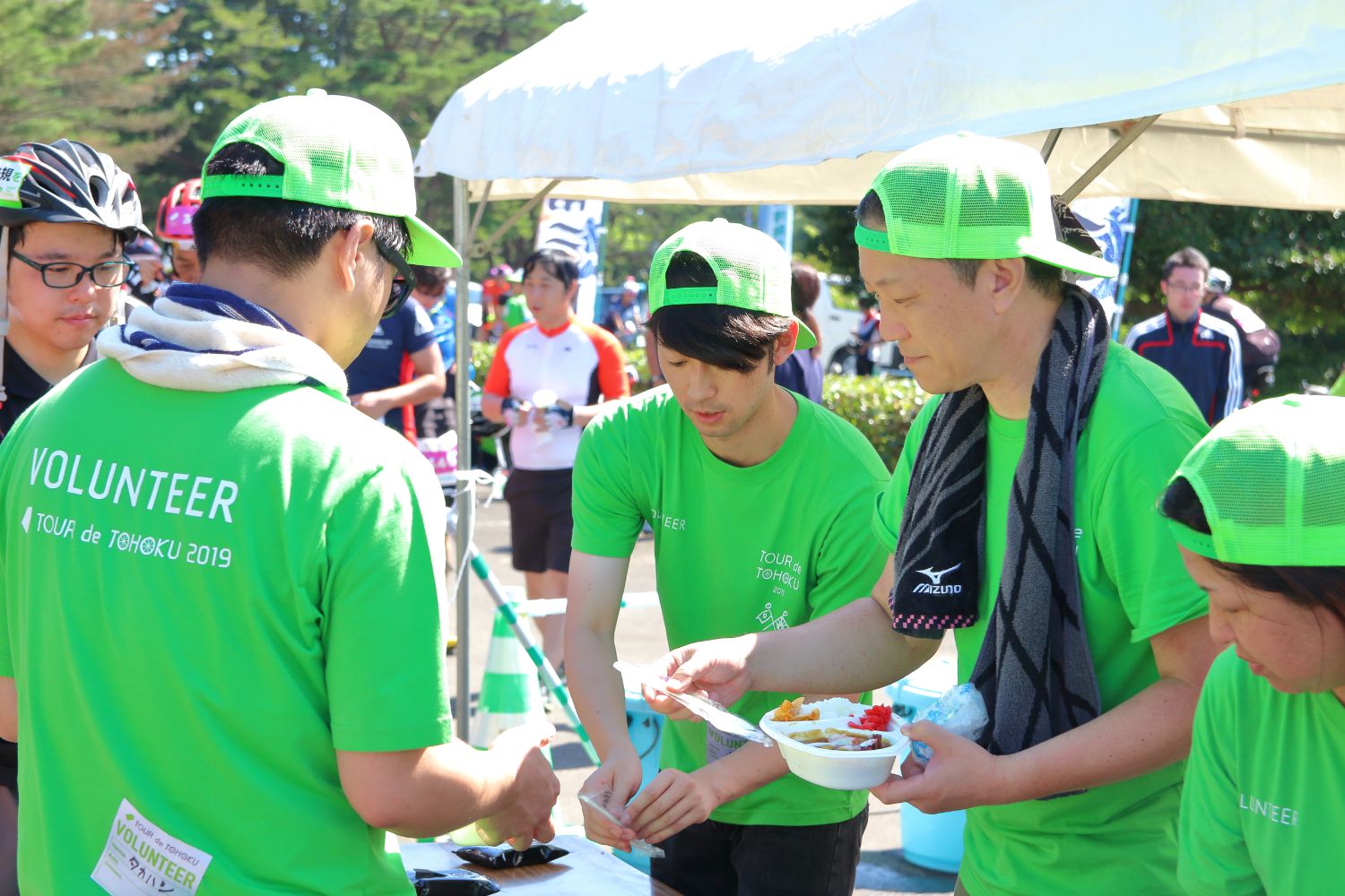 お揃いの黄緑色のキャップを被りシャツを着たボランティアの方々が、ライダーにカレーライスを手渡そうとしている写真