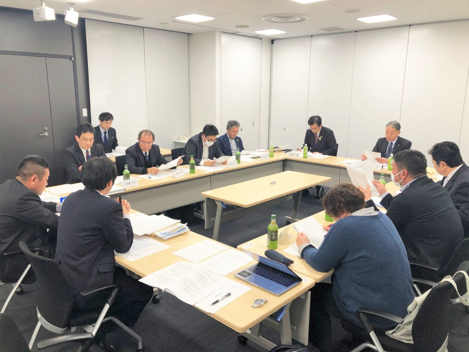 会議室でロの字型に設置された長机を囲んだスーツを着た男性たちが、資料を読みながら真剣に会議をしている写真