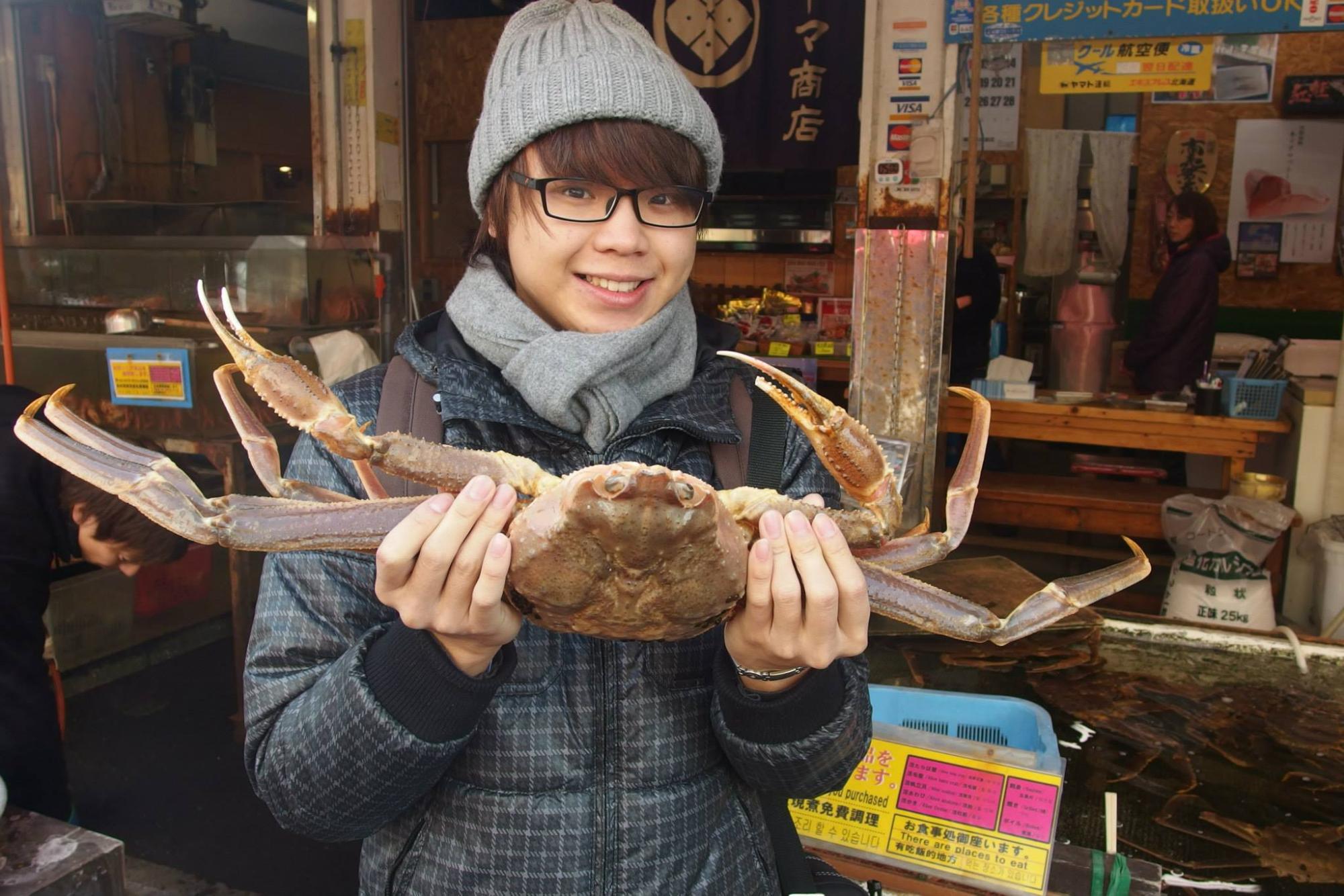 沢山のカニが入った水槽のある店内で、灰色のニット帽とマフラーを着けた陳さんが両手で大きな生きたカニを持ち上げ、笑顔でカメラを見ている写真