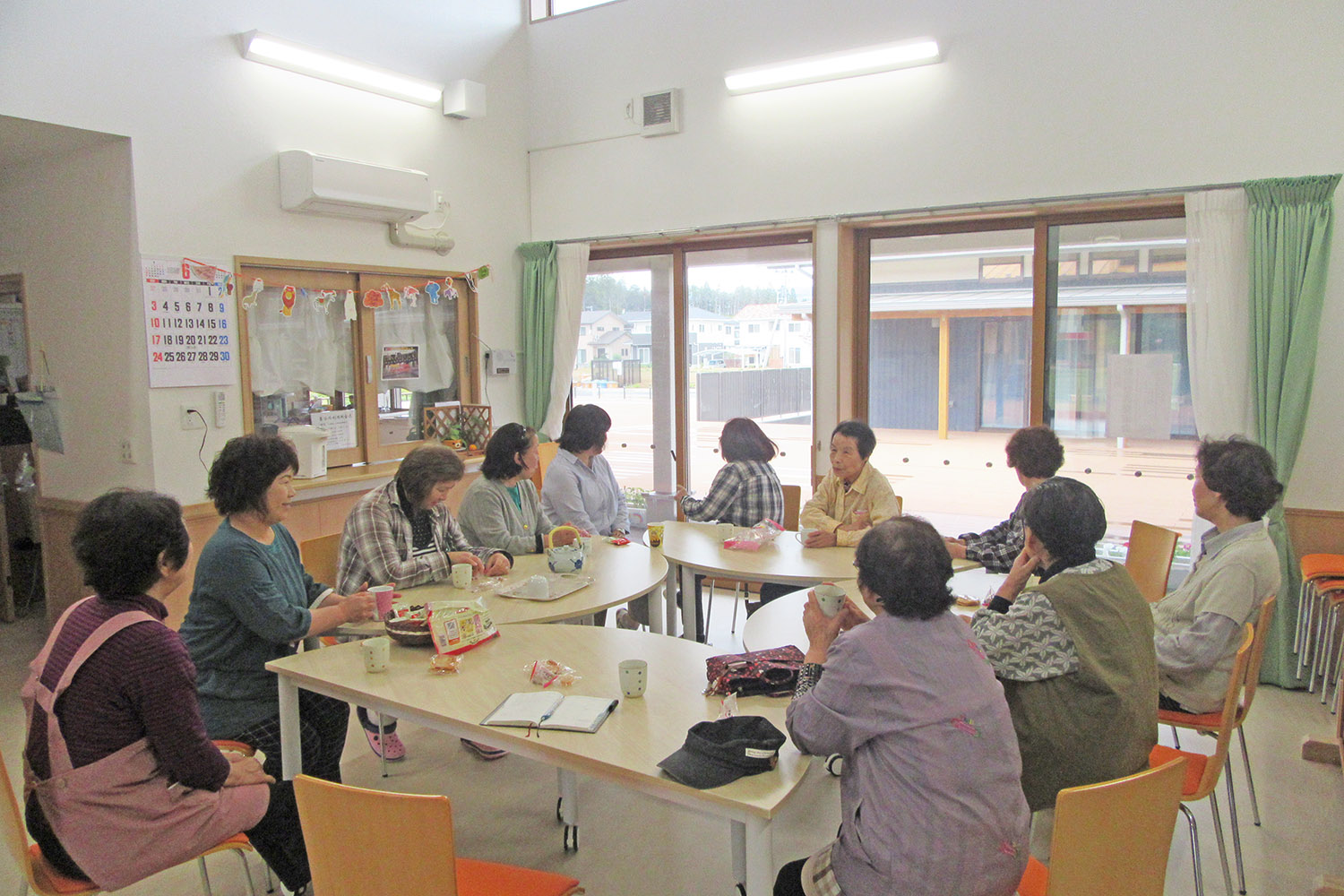 室内のテーブルを囲んで11人の高齢の女性が座ってお茶やお菓子を楽しみ、大きな窓の外を眺めながら談笑している写真