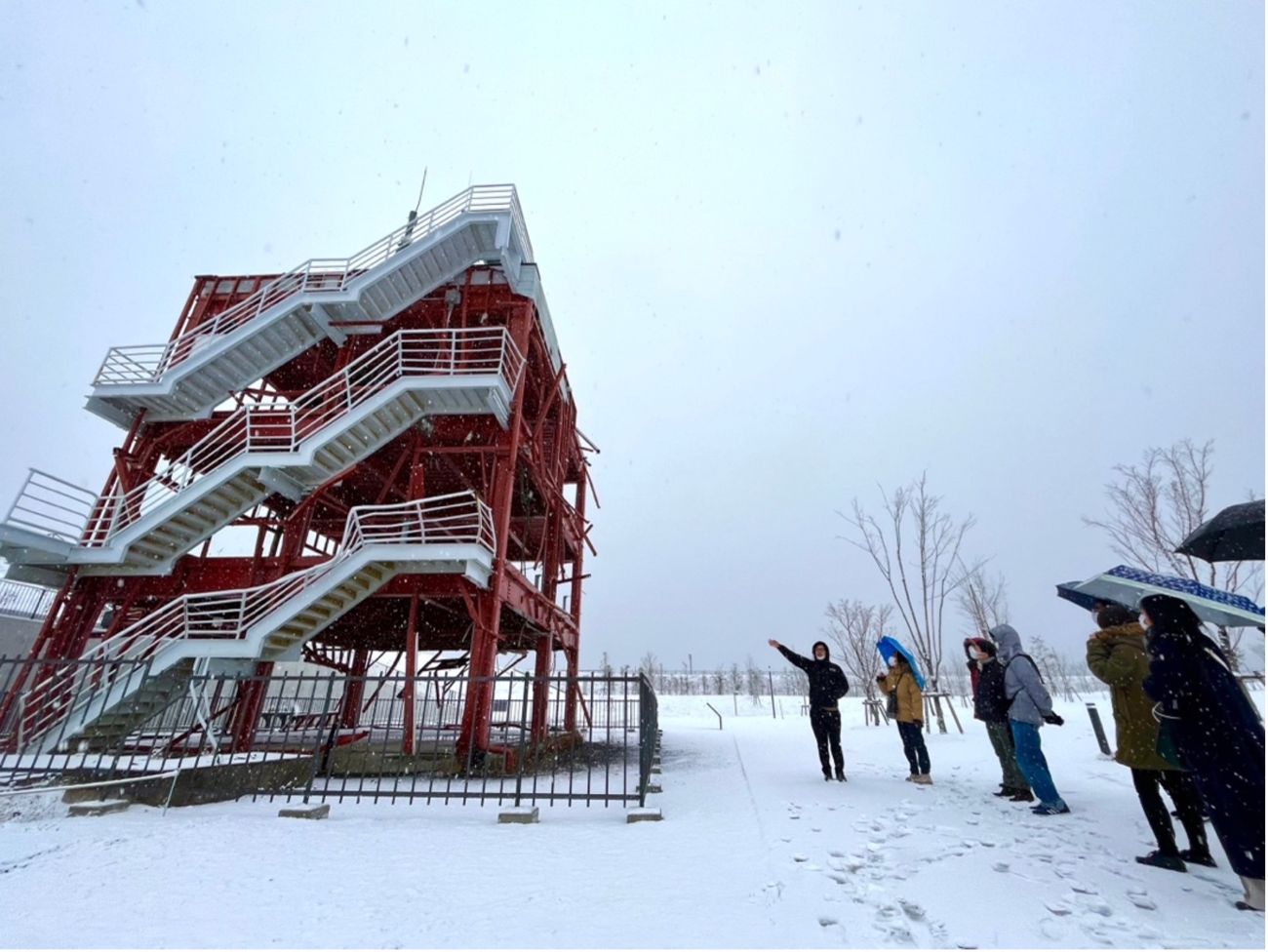 雪の積もった屋外で若者たちが、東日本大震災で津波に耐えた赤い鉄骨の建物を見上げながら説明を聞いている写真