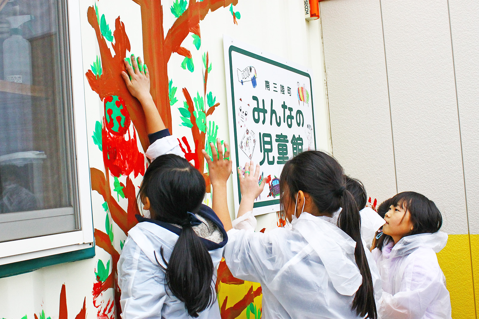 白いビニールのレインコートを着た子どもたちが外壁に書かれた木の絵に緑色の絵の具をつけた手で手形を押している写真
