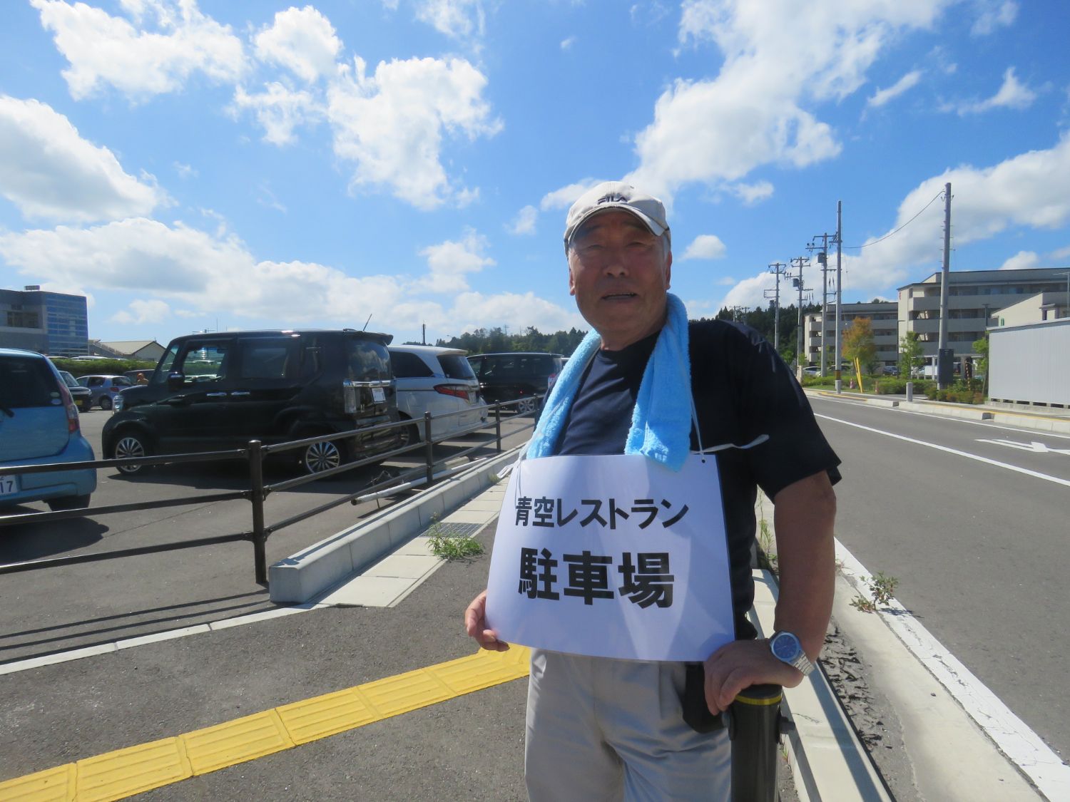 晴れ渡る青空の下、駐車場の一角で「青空レストラン 駐車場」と書かれた紙を首から下げた高齢の男性が、帽子と青いタオルを身に着けて立ち、来場者の案内をしている様子が写された写真