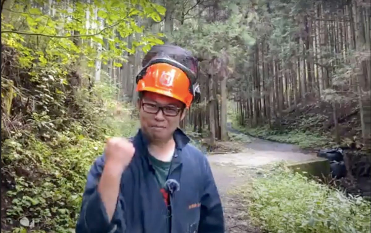 森林の中の細い舗装道路のわきに立つ、オレンジ色のヘルメットと作業服を身につけた男性が、右手を握ってガッツポーズをして笑顔を見せている写真