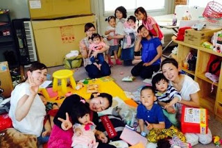 室内の畳やカーペットの上に、子ども用のおもちゃや絵本が広げら中央付近には床に座っている母親と子どもたち、左前方では、女性2人がピースサインをしてカメラに笑顔を向け子どもたちも一緒に手を振っている室内でくつろぐ親子たちを写した写真