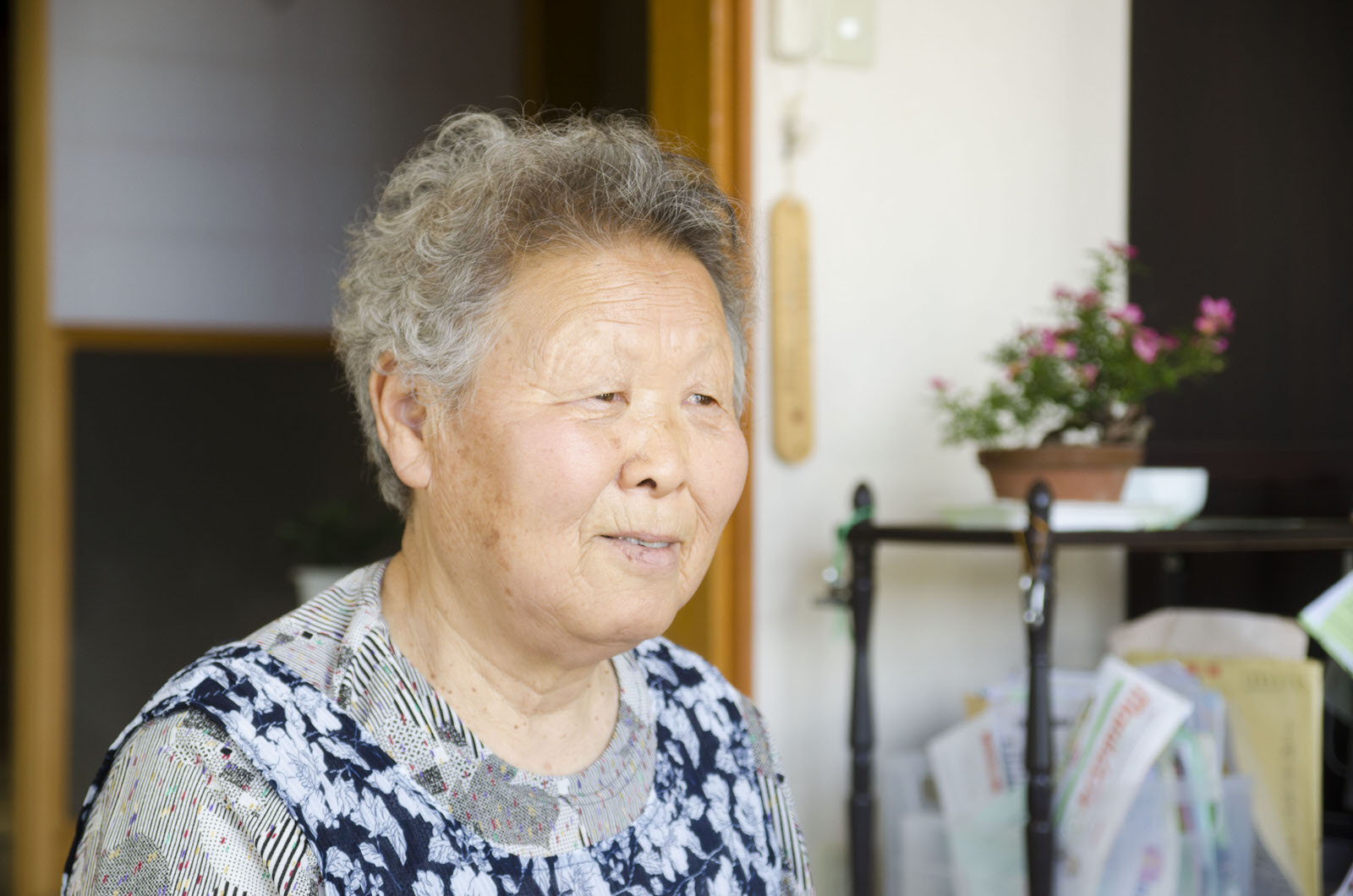 鉢植えや日用品が置かれている室内で、穏やかな表情で話している様子をとらえた首藤さんをアップで撮影した写真