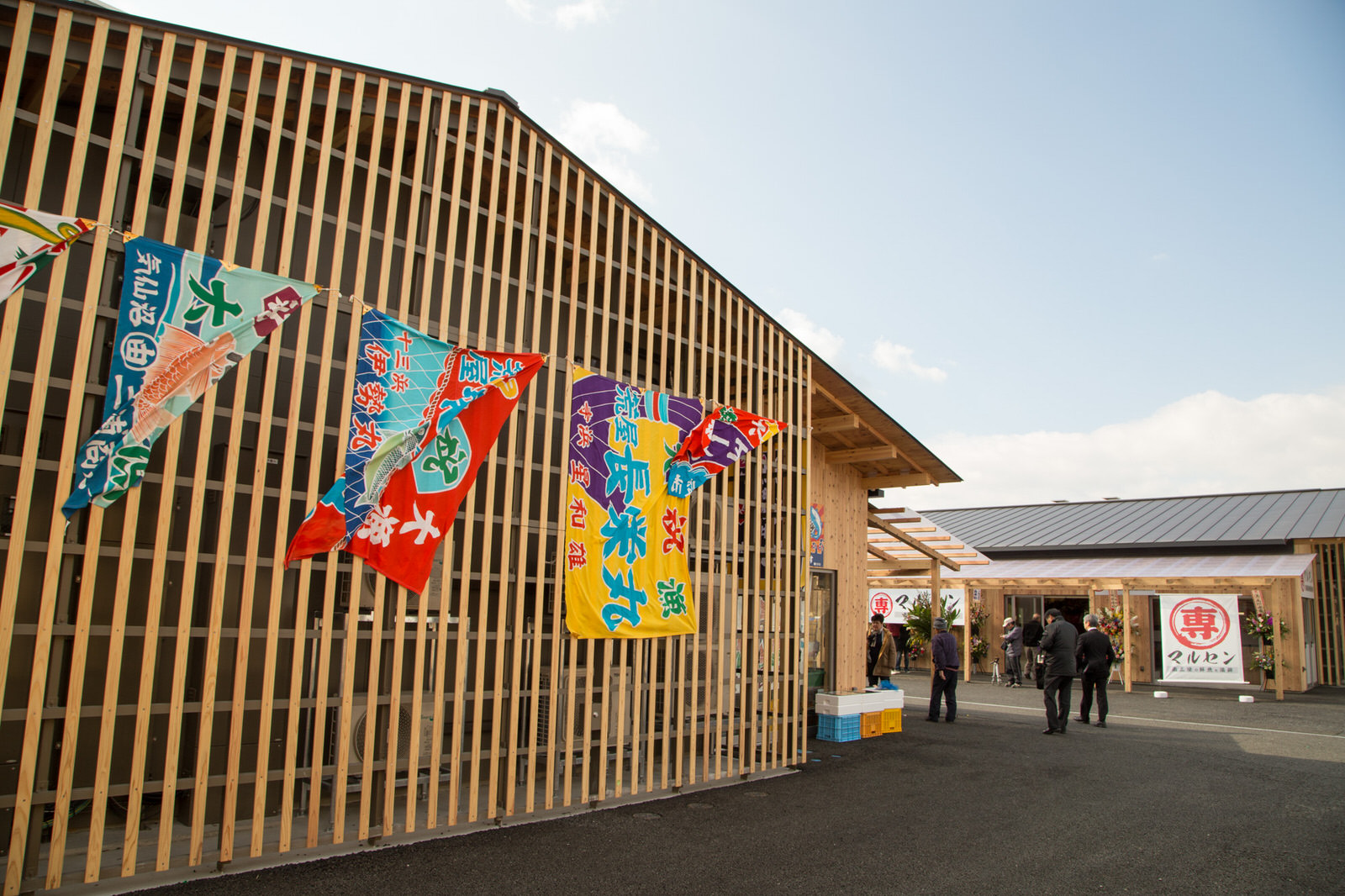 木の格子が印象的な建物の壁に、色鮮やかな大漁旗が複数掲げられていて、その隣にある別の建物の入口には「マルセン」と書かれた白い大きな旗が見える、店舗の外観の写真