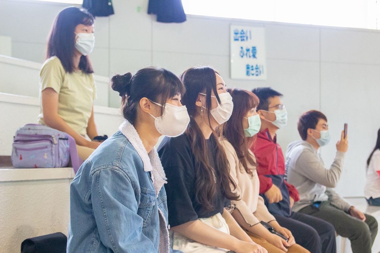 マスクをつけた参加者たちが室内の観客席に座り、前方を見つめている様子の写真