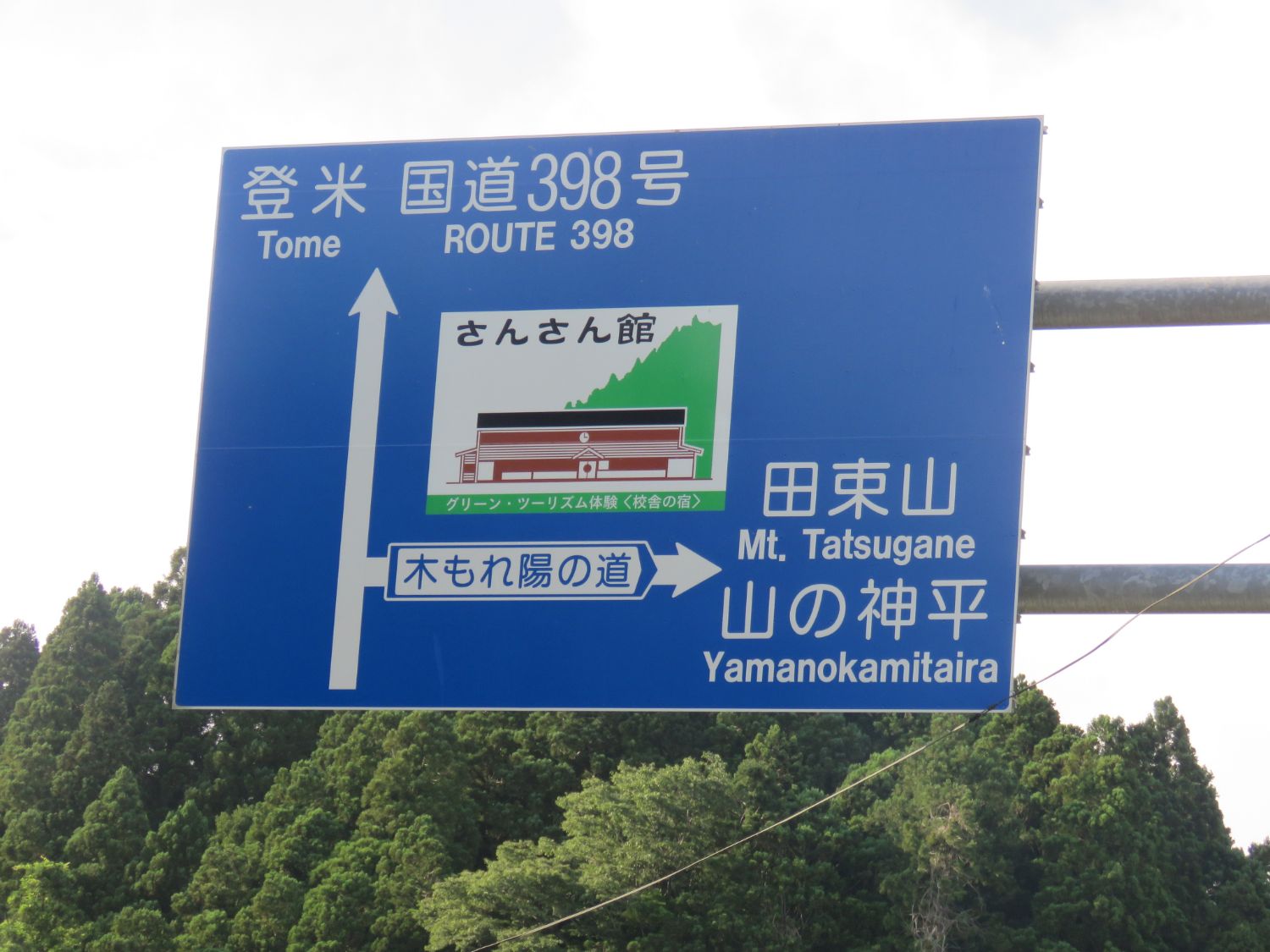 青い道路標識に「登米」「国道398号」や右側の木もれ陽の道方面に「田束山」「山の神平」などの文字とさんさん館の建物のイラストが描かれた道路案内標識の写真