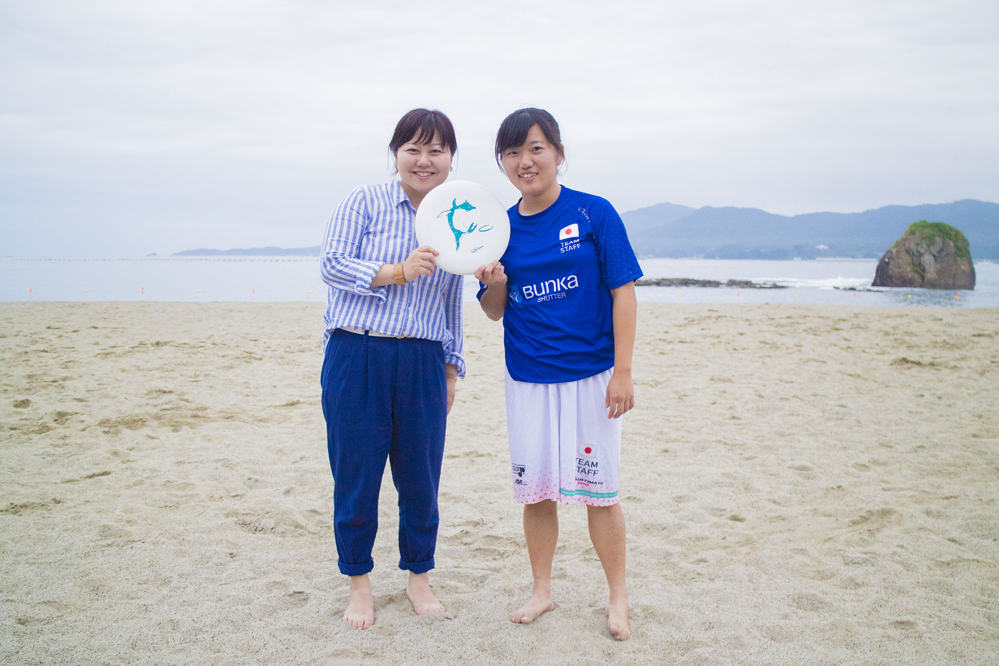 砂浜で海を背景に、ストライプのシャツと青いズボン姿のリポーターの女性と、青いユニフォームを着た宮岡さんが、裸足でフリスビーを持って笑顔で立っている写真