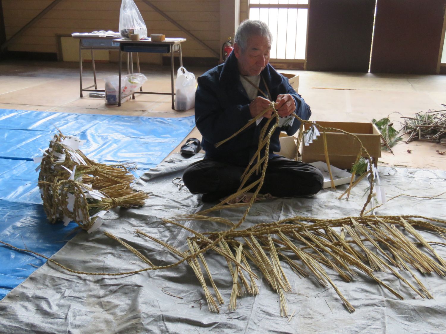 ブルーシートの上にしめ縄が重ねて置かれ、あぐらを組んで床に座りしめ縄を編んでいる佐藤和行さんの写真