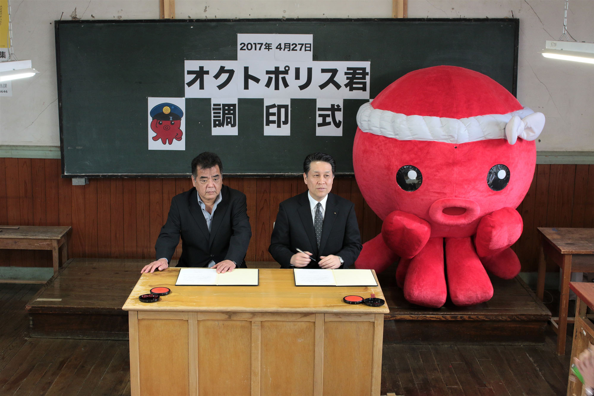 「2017年4月27日 オクトポリス君 調印式」と書かれた黒板の前で、赤いタコのキャラクター「オクトポリス君」とともにスーツ姿の2人の男性が調印を行っている写真