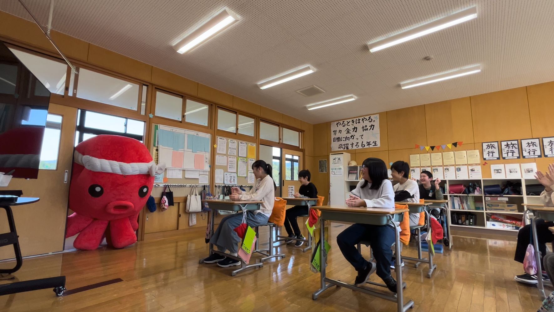 教室のドアから赤いたこのオクトパス君のマスコットが入ってくるのを、机に座った子どもたちが笑顔で迎えいる様子の写真