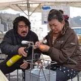 隣に座っている村山さんが、茶色の上着を着た女性参加者が両手で持った棒の先にあるドロドロになったオレンジ色のガラスの元をバーナーで熱を加え、女性が棒を回しているガラス玉作り体験の様子の写真