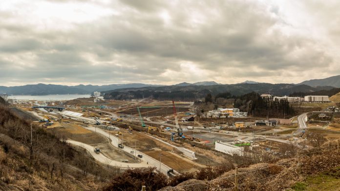 現場には複数のクレーンや建設車両が見られ、建物や道路が部分的に建設され背景には丘や山が広がり、空が曇っている建設現場の写真