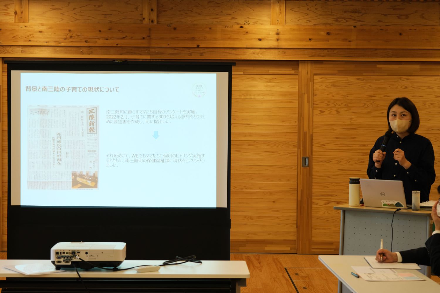 マスクを着けた栗林さんがマイクを持って立ち、スクリーンに表示されたスライドをもとに説明をしている写真