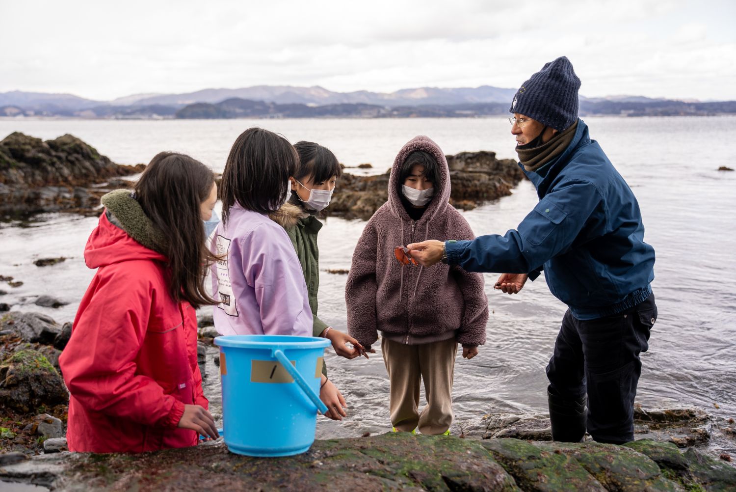 海辺の岩場で、南三陸少年少女自然調査隊の子どもたちがバケツを持ちながら関係者の男性の説明を真剣に聞き、海の生きものを観察している様子の写真