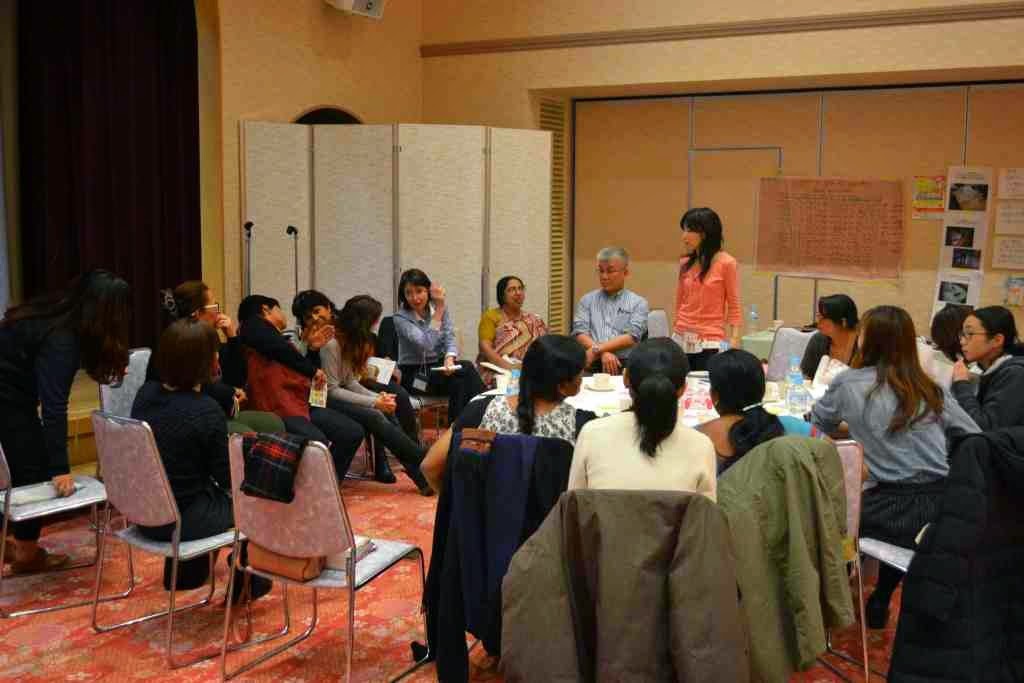 部屋の中央にはテーブルがあり、その周りに人々が集まりテーブルの上には飲み物や書類などが置かれており、右側には立って話している女性がいる勉強会での写真