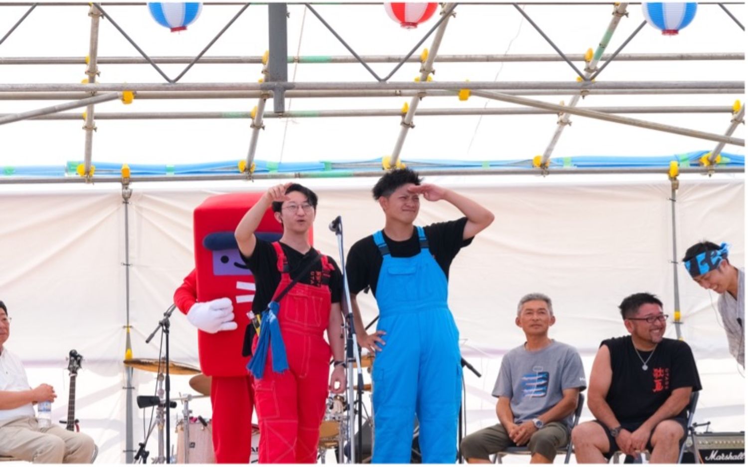 舞台の上で赤いオーバーオールの男性と青いオーバーオールの男性が指をさしたり敬礼するポーズをとり、後ろに赤いキャラクターの着ぐるみが控えている様子の写真