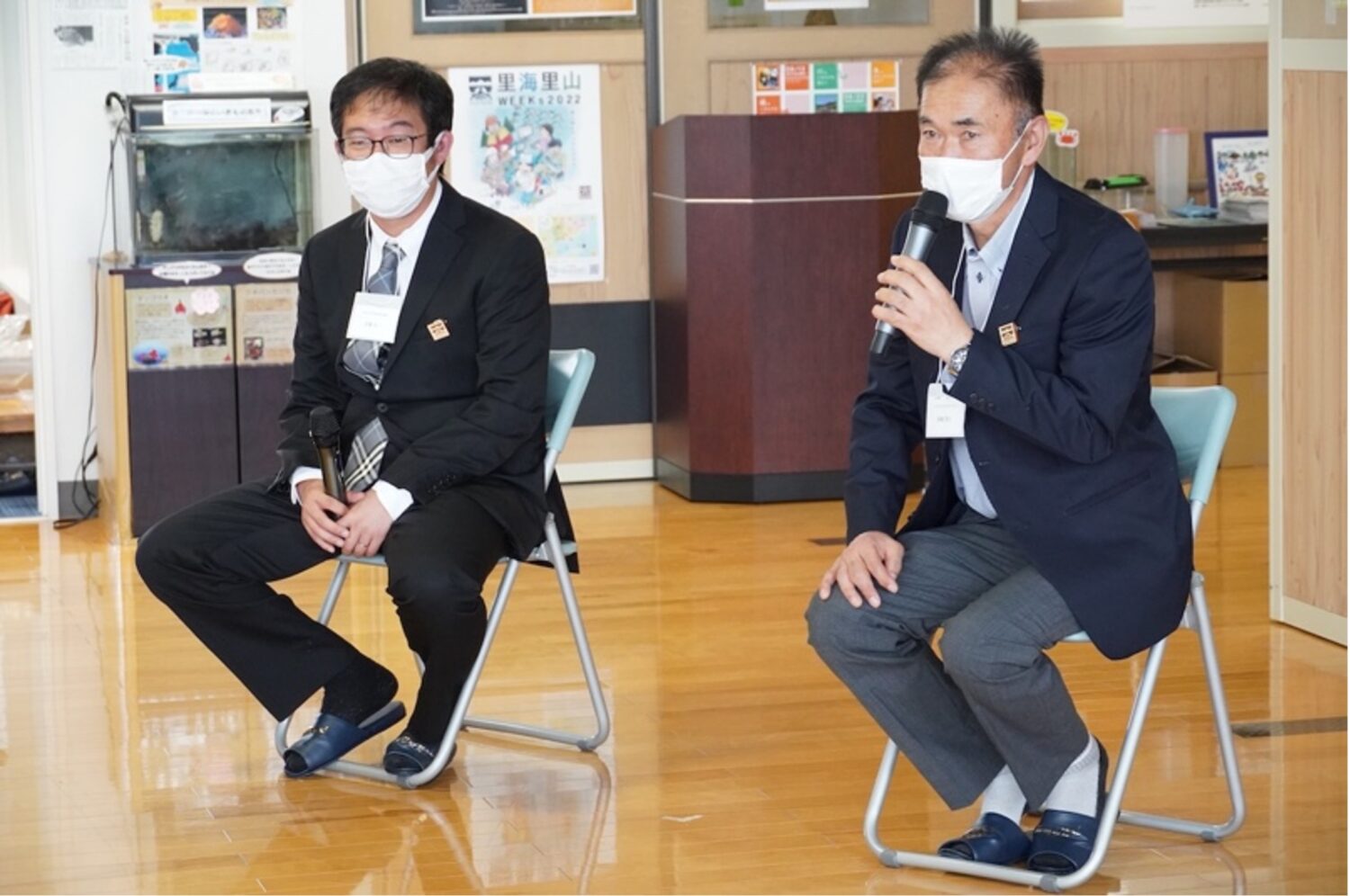 スーツ姿の関係者が2人椅子に座っており、右の後藤清広さんがマイクを持って話をする様子の写真