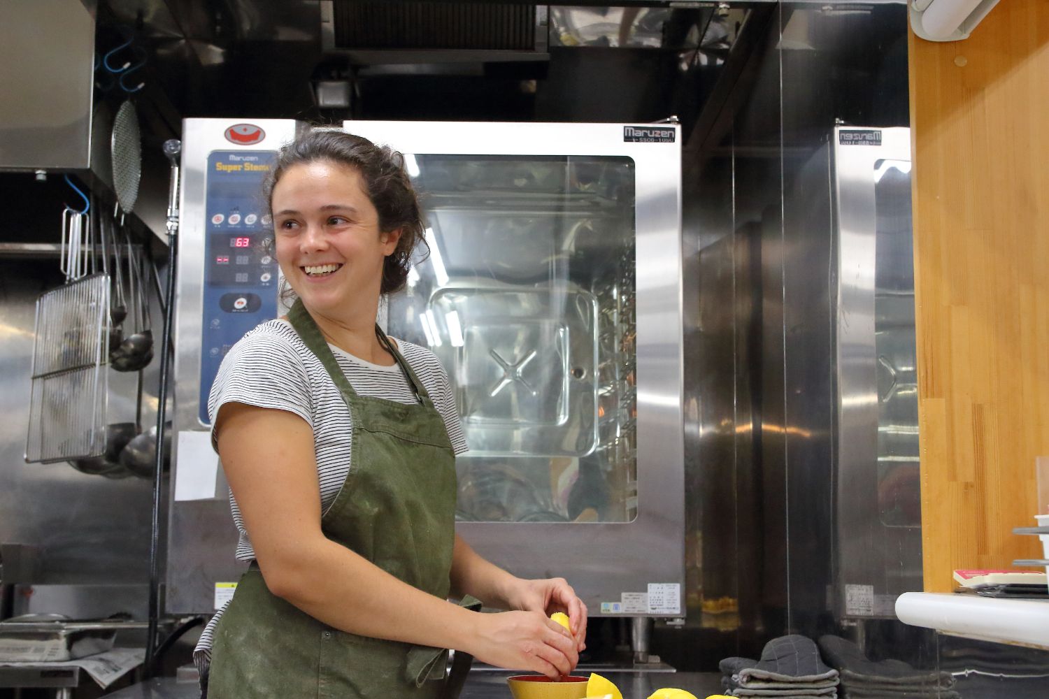 モスグリーン色のエプロンを着用したディアナ・ピサノさんが、料理をしながら笑顔で後ろを振り返っている写真