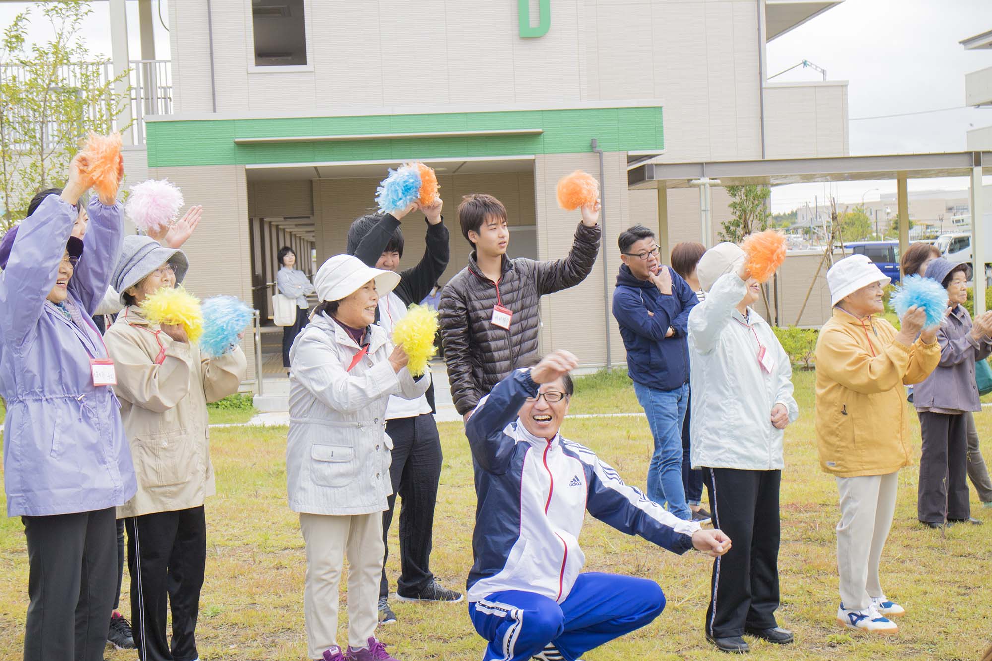 カラフルなポンポンを持った高齢者や若者たちが笑顔で手を振ったり拍手をしたりしており、前方ではジャージ姿の男性がしゃがんで手を上げて笑顔で応援している写真