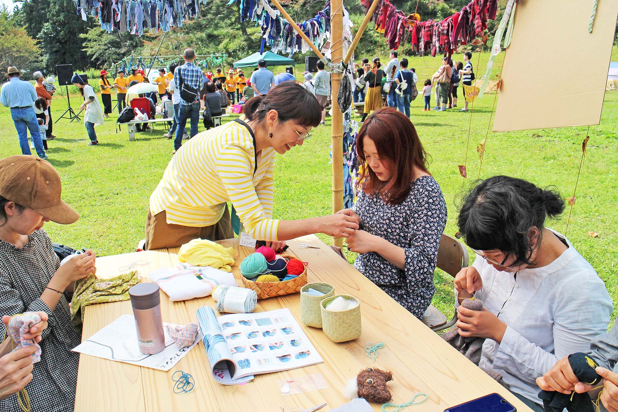 テーブルの上に毛糸や手芸道具を並べて屋外で開かれたワークショップで、スタッフが参加者に手芸のやり方を優しく教えている様子の写真