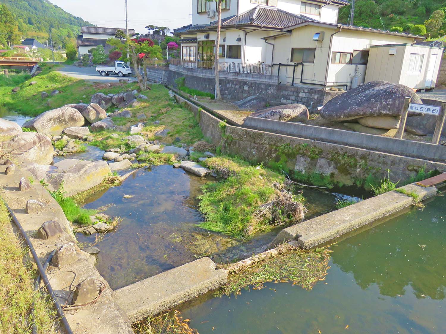 川沿いにある民家の前に重ね石が置かれており、周囲には自然の岩や水路があり、奥には橋がかかっている風景写真