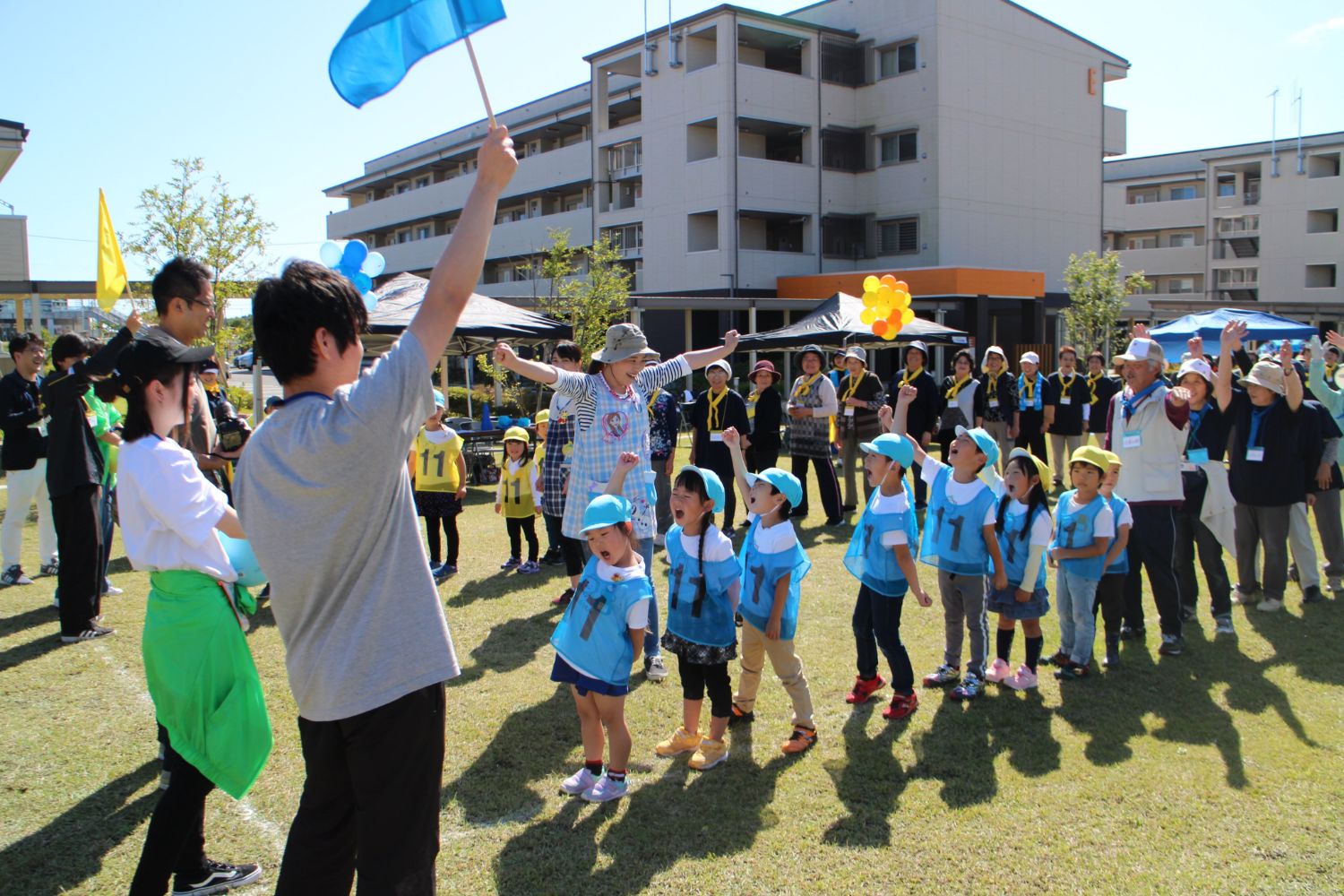 青い旗を持った大人が水色のビブスを着た幼稚園の子供たちを先導している様子