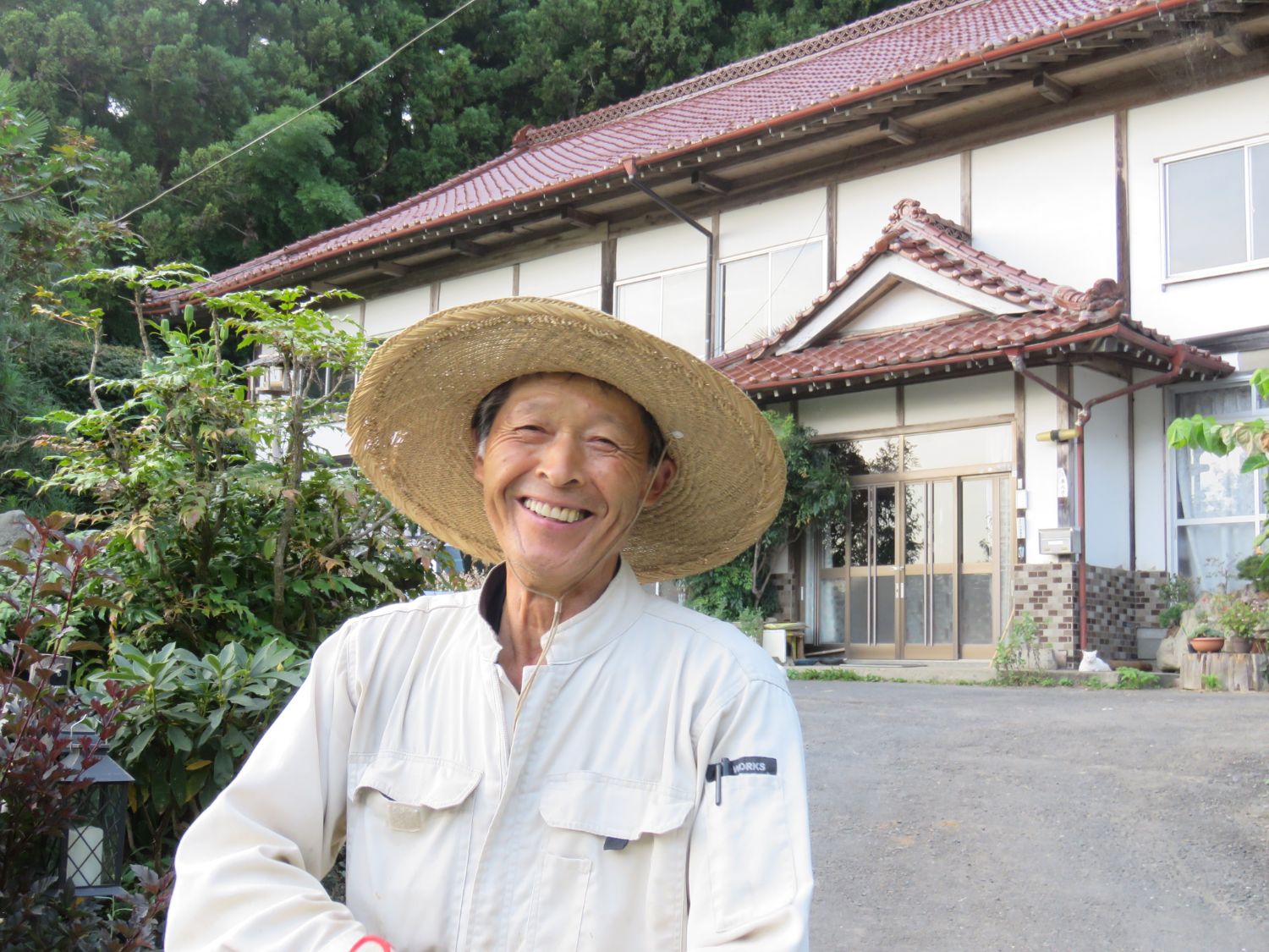 農村の家の前で麦わら帽子をかぶった男性が笑顔を見せている写真