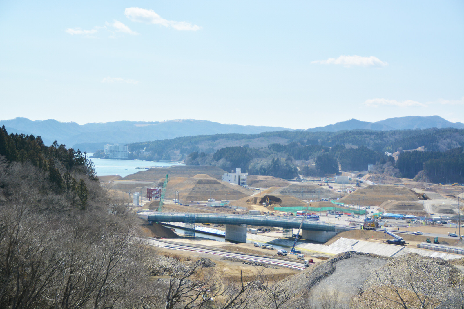 海と山に囲まれた平地で大規模な造成工事が進んでおり、新しい道路や橋が建設中の様子を高台から撮影した写真
